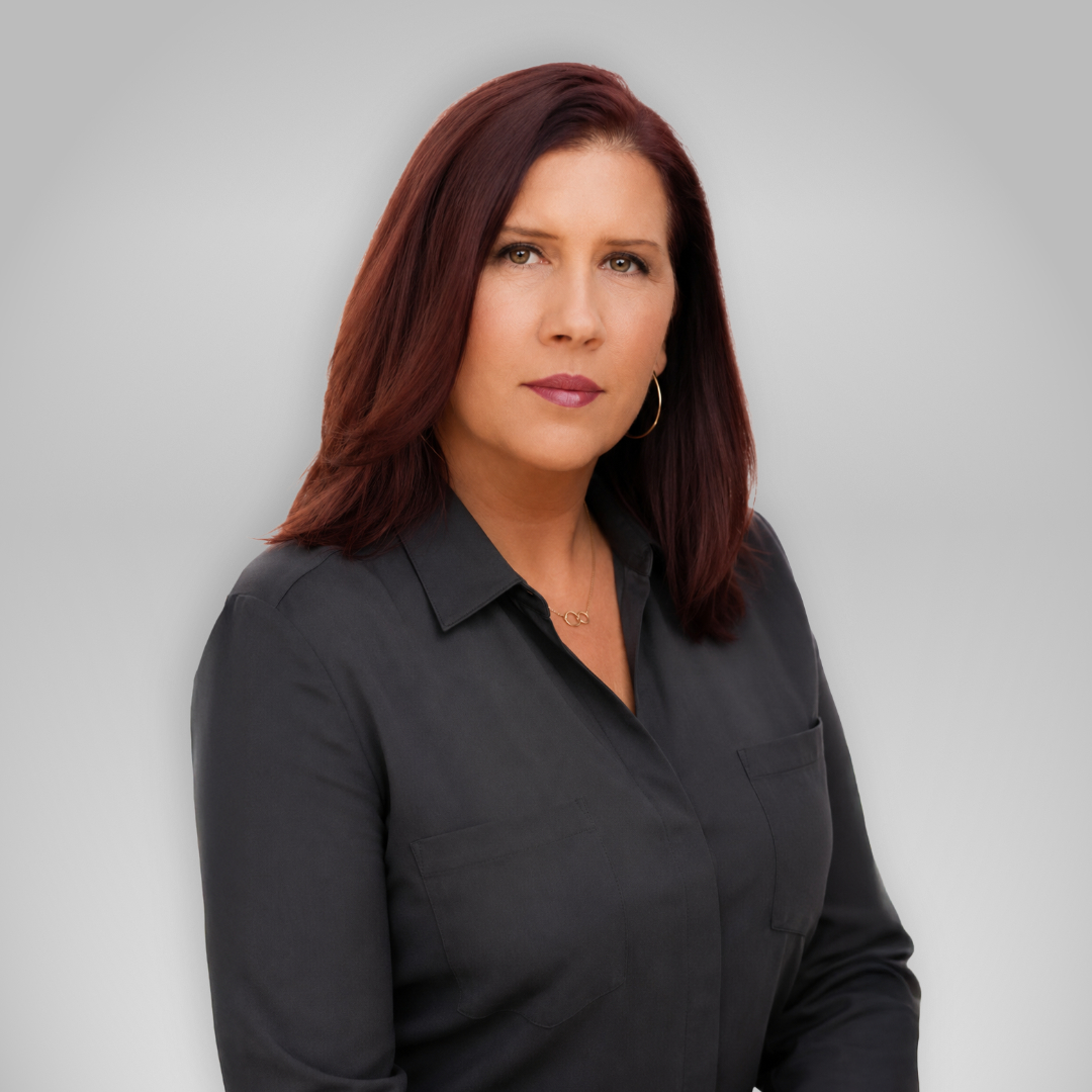 Professional headshot of a woman with shoulder-length auburn hair wearing a dark gray blouse, neutral expression, and a light gray backdrop.